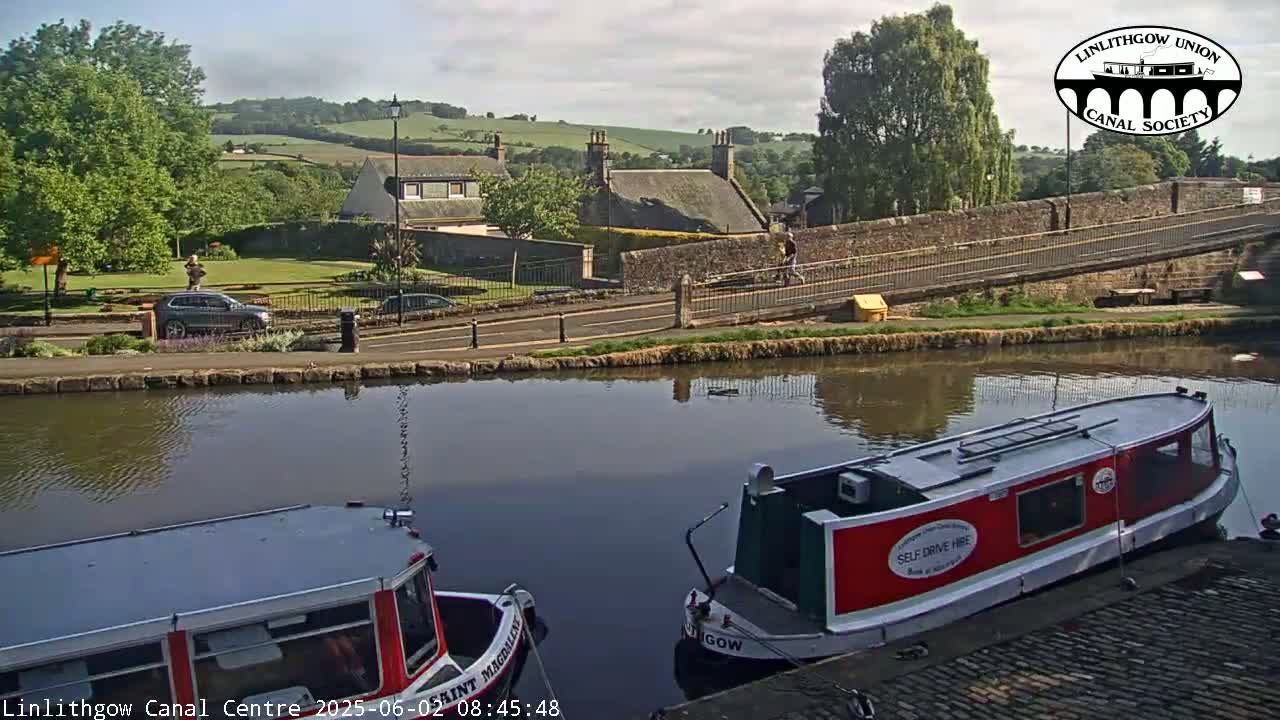 Two narrowboats are moored on a calm canal, with a stone bridge and buildings visible in the background under a partly sunny sky.