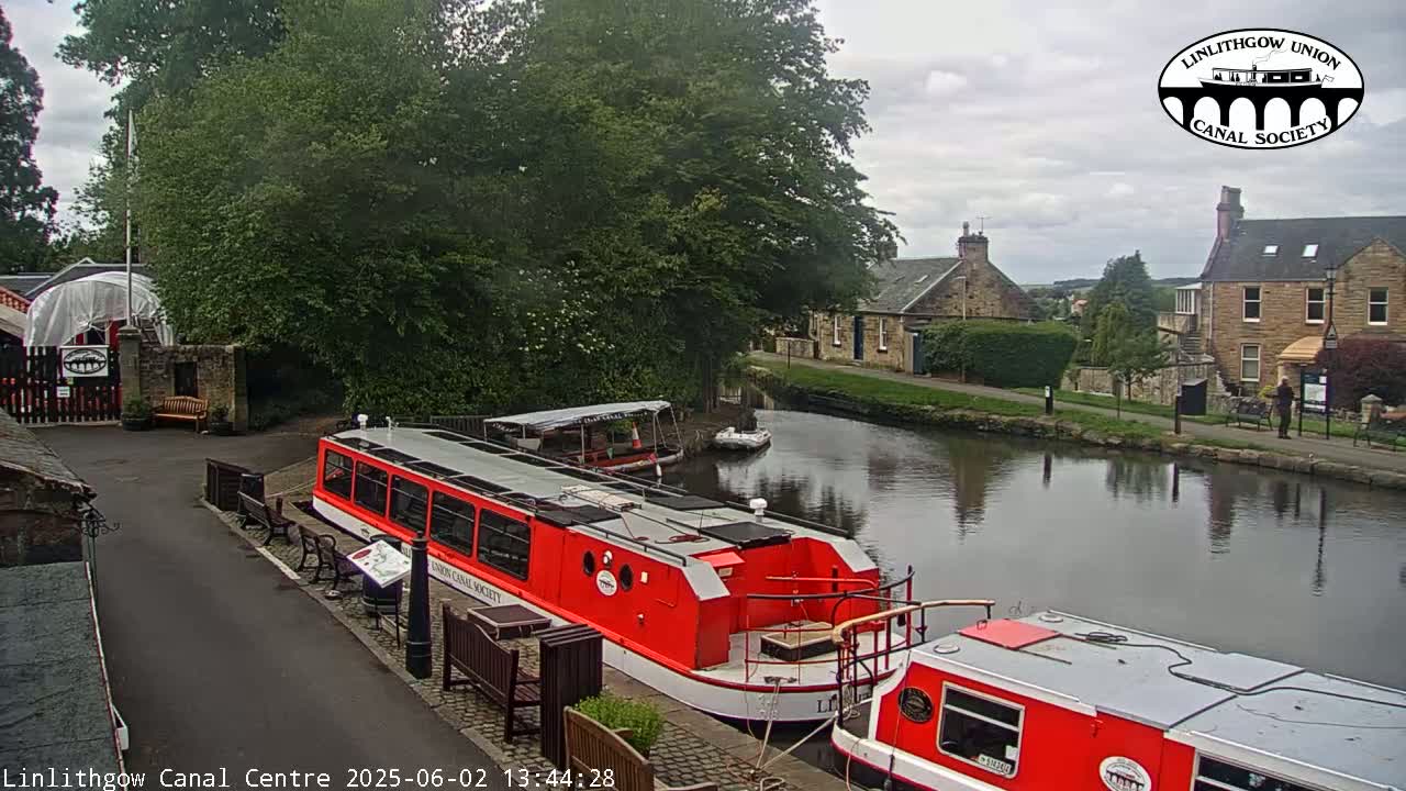 Two red canal boats are docked near a stone building and lush green trees under an overcast sky.
