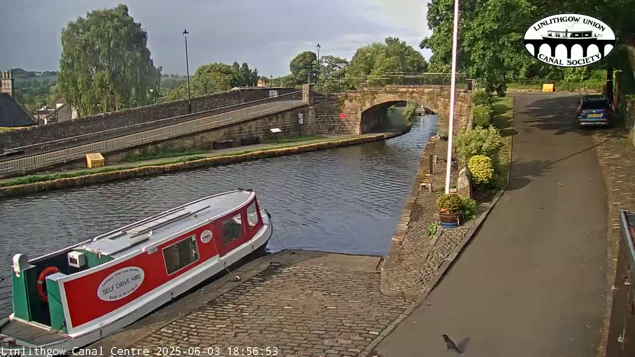 A red narrowboat is moored at a canal lock under a partly cloudy sky, with a stone bridge and roadway visible in the background.