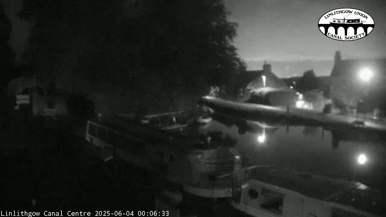 A nighttime, low-light view shows several boats moored along a canal, with buildings and trees visible on the banks.
