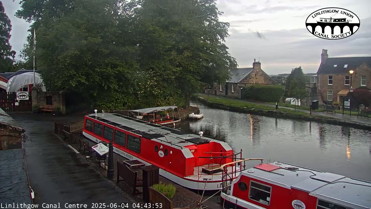 Two red narrowboats are moored alongside a canal, under an overcast sky, next to buildings and lush green trees.