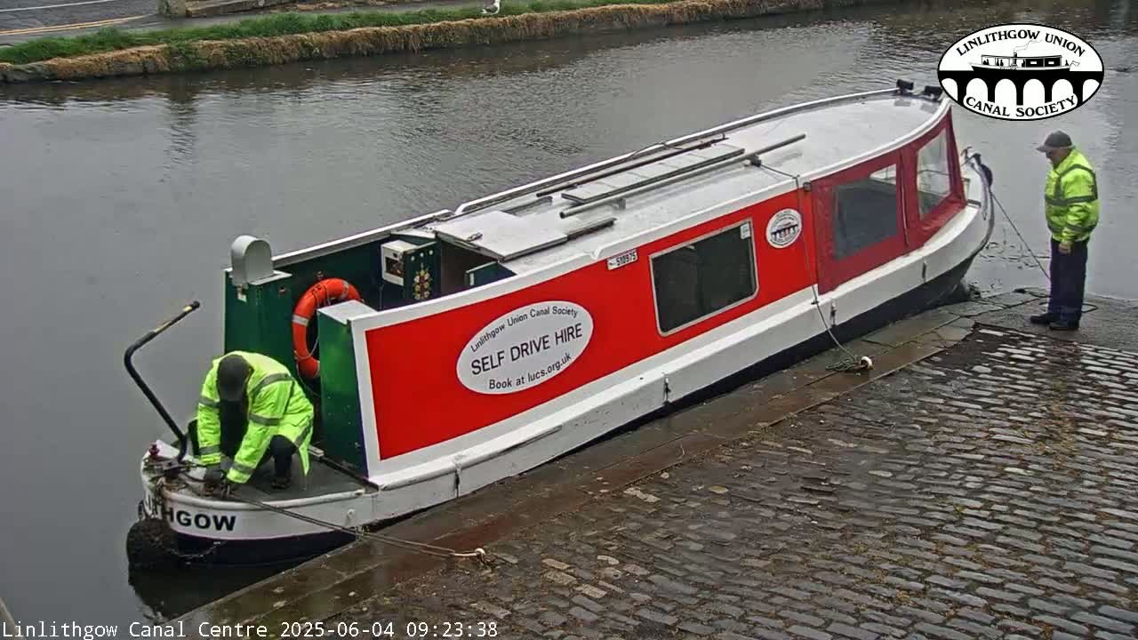 A red and white canal boat is moored at a cobblestone dock beside a calm canal, with two people working near the boat's bow.