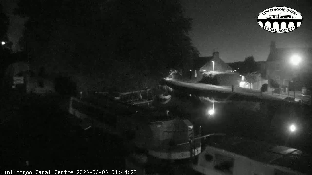 A nighttime, low-light view of a canal with several boats moored alongside a building under a partly cloudy sky.