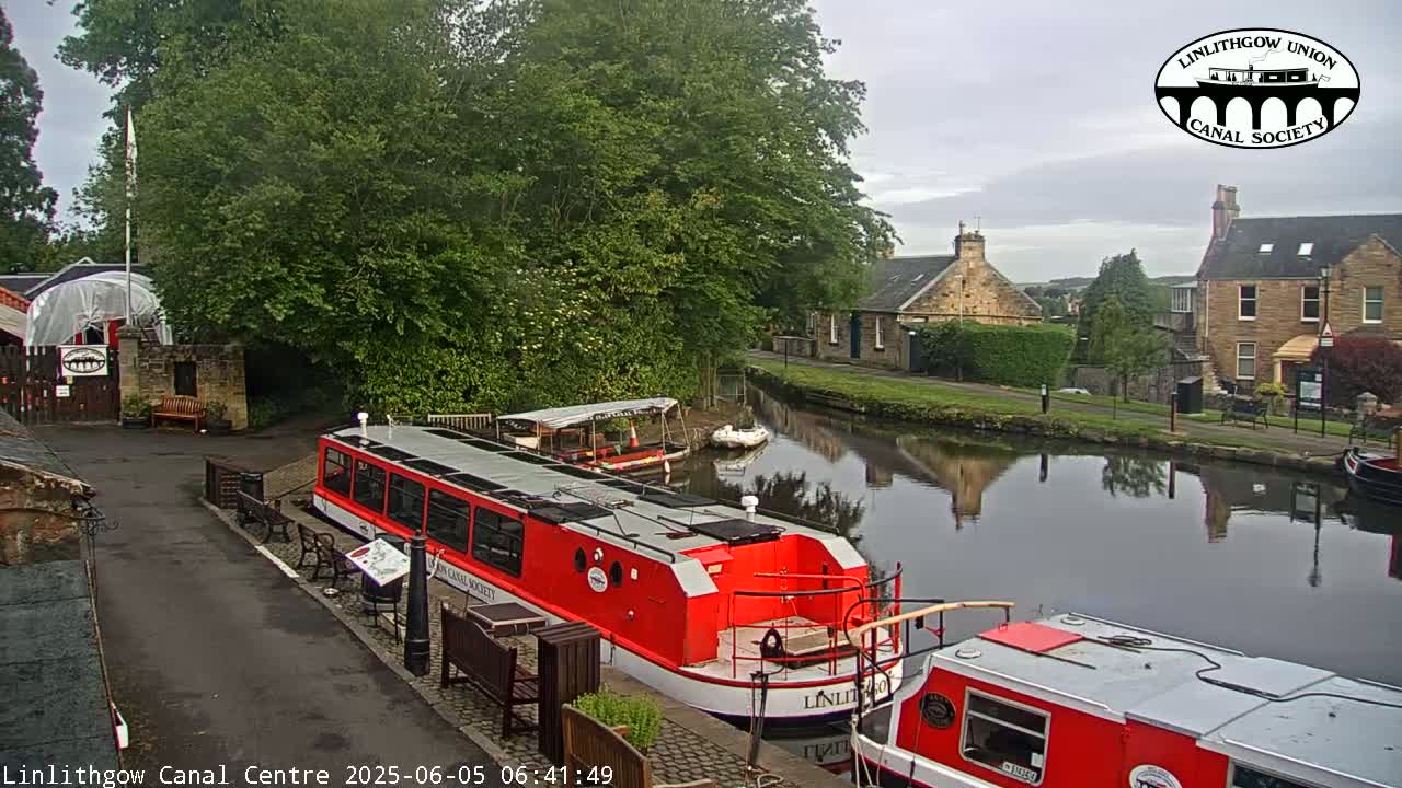 Two red canal boats are moored near a canal side, with stone buildings and lush green trees in the background under an overcast sky.