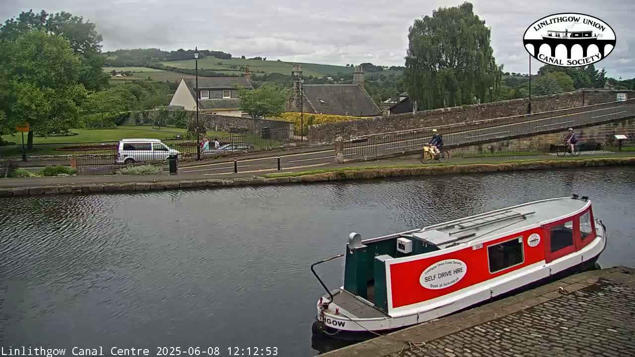 A red and white narrowboat is moored on a canal beside a paved walkway; overcast skies are visible above a quiet residential area with a few people walking and cycling.