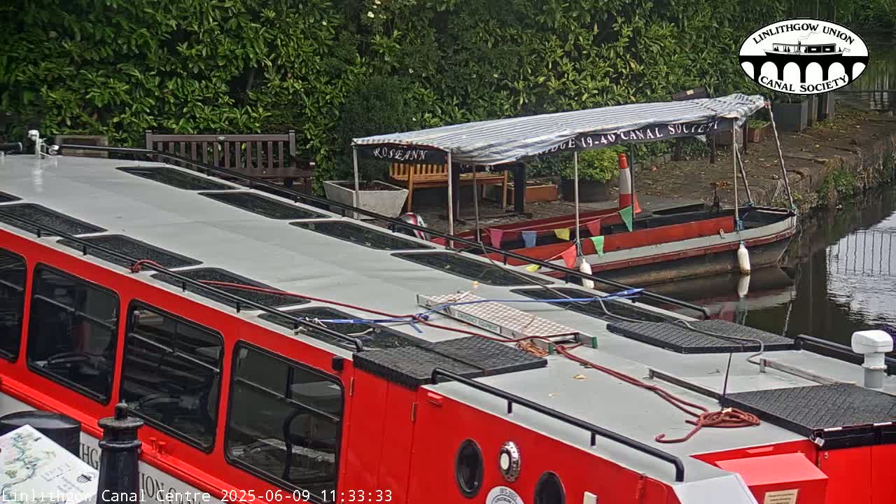 A red passenger boat is docked next to a smaller, red and white boat on a canal under overcast skies.