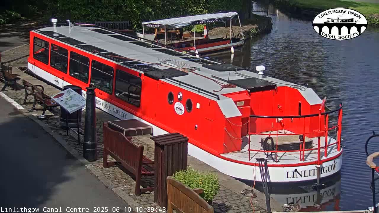 A red and white canal boat is docked next to a smaller, gray boat on a sunny day.
