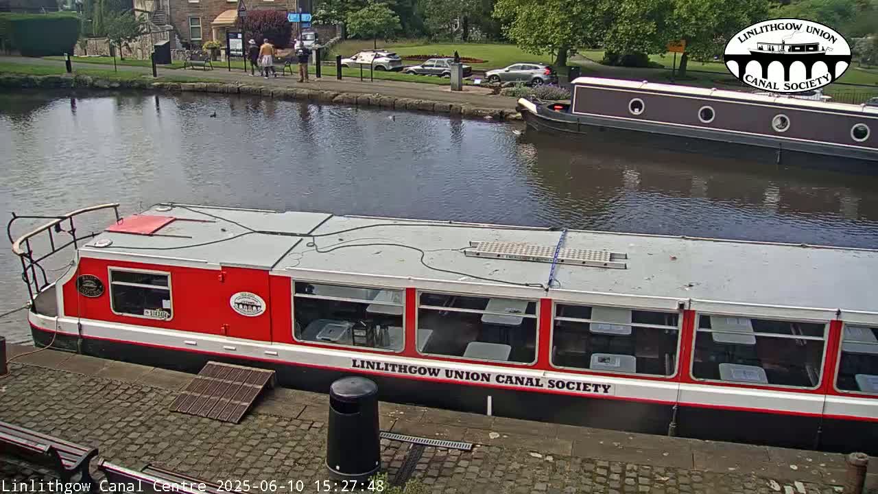 A red and white canal boat is docked next to a dark brown canal boat on a calm canal, under a sunny sky.