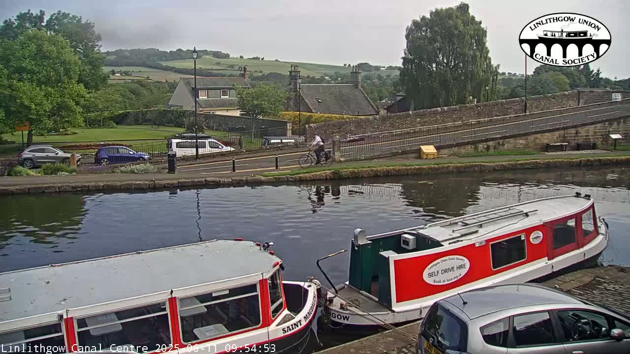 Two canal boats are moored by a canal side, with a person cycling past a stone bridge and parked cars in the background under an overcast sky.