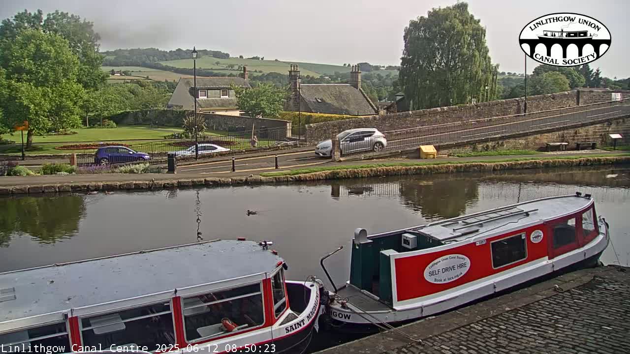 Two narrowboats are moored on a canal beside a stone bridge and road, with cars parked nearby, under a hazy sky.