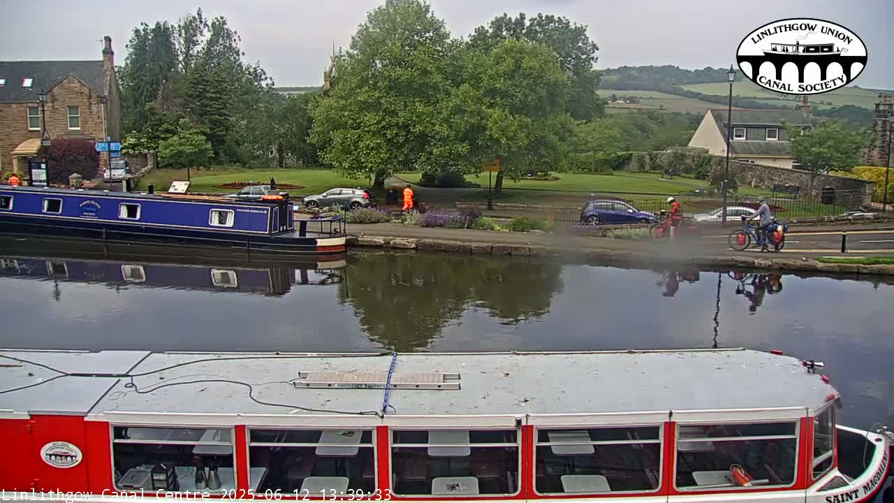 A canal scene on an overcast day shows a red and white passenger boat in the foreground, a blue narrowboat moored further away, cars parked along the canal bank, and two cyclists riding along a path.