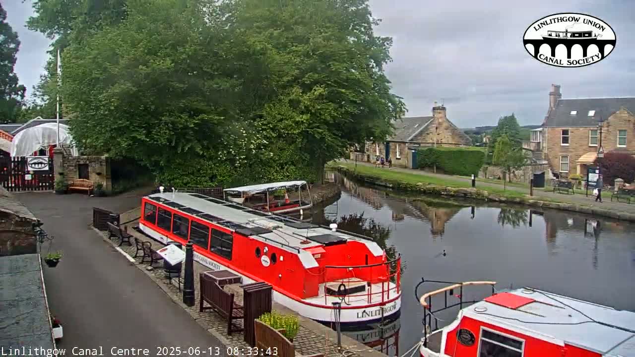 A red canal boat is docked near a canal-side building, with another boat visible further down the waterway under an overcast sky.