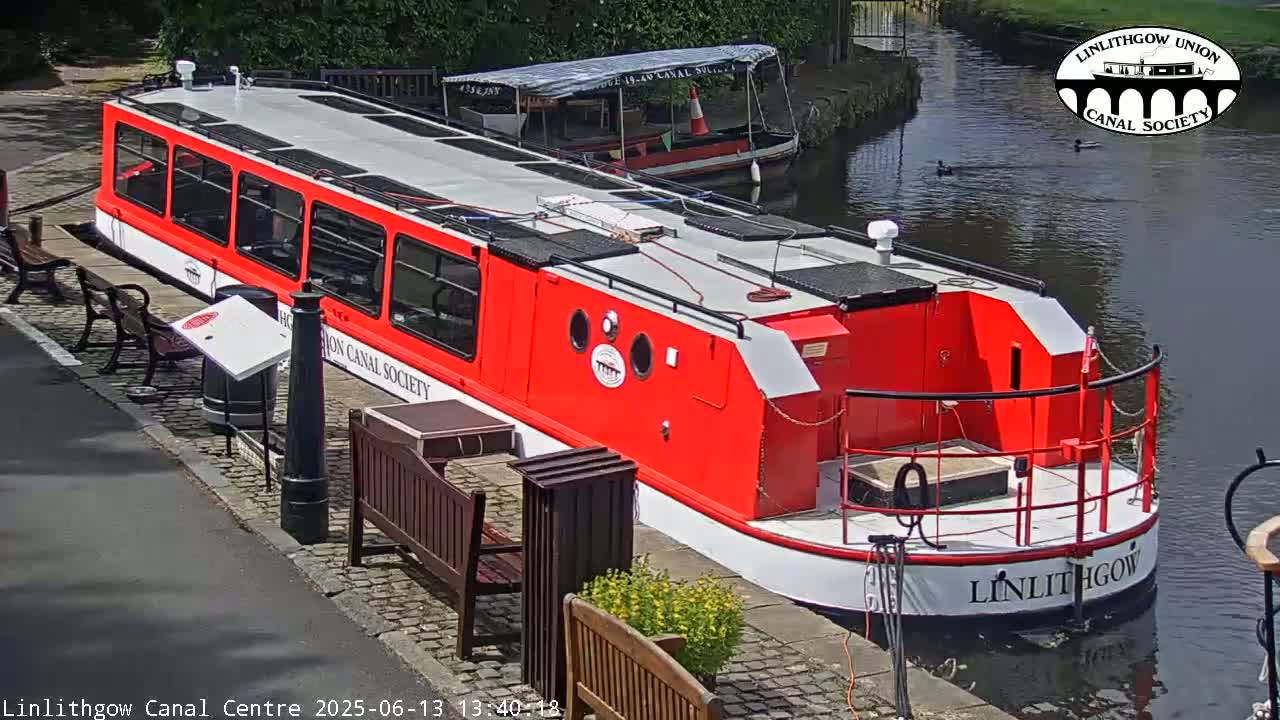 A red and white canal boat is moored alongside a paved walkway, with benches and small plants visible nearby, on a sunny day.