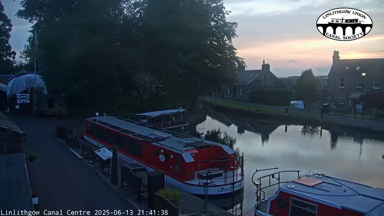 A red canal boat is docked near a calm canal reflecting a twilight sky, with buildings and trees lining the waterway.