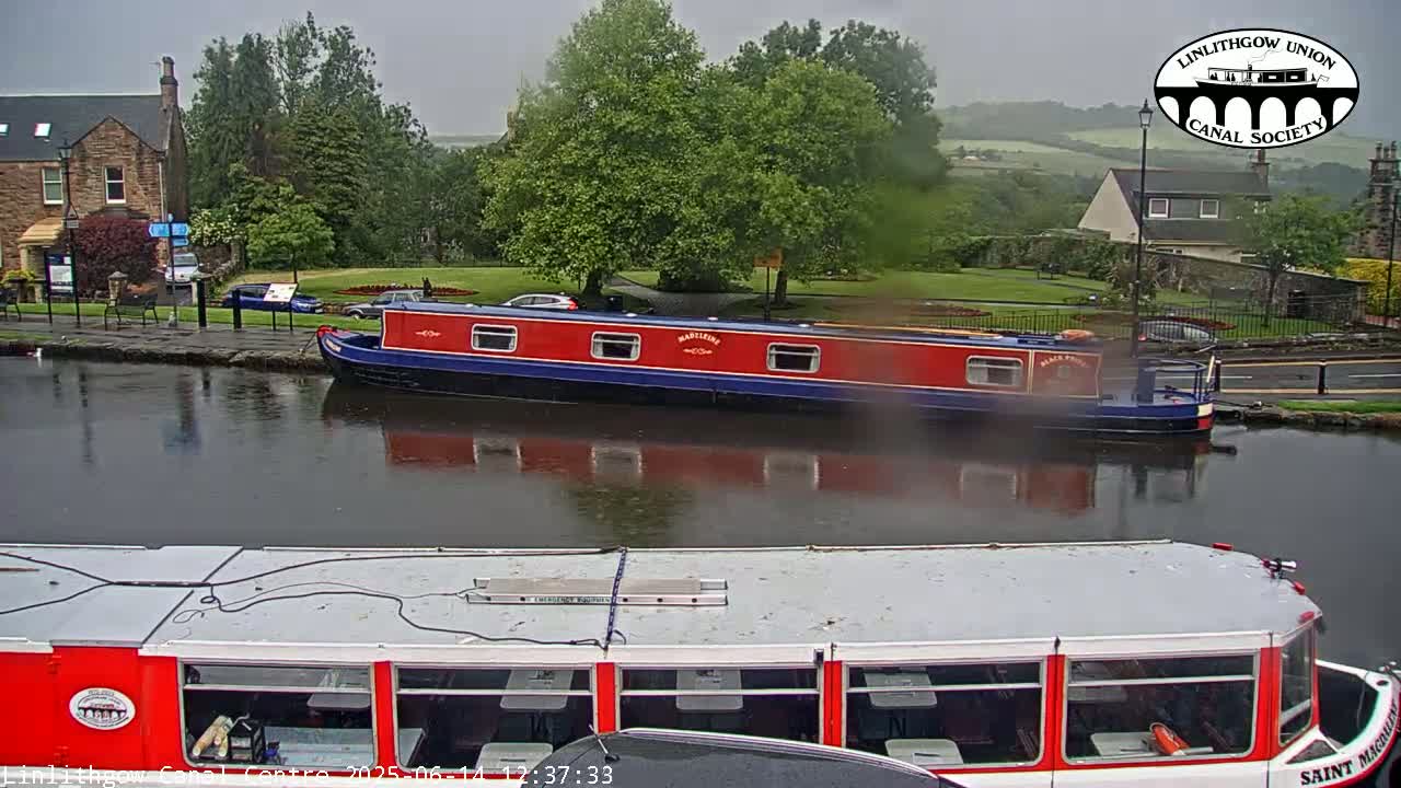 A red and blue narrowboat is moored on a canal under an overcast sky, with another boat visible in the foreground.