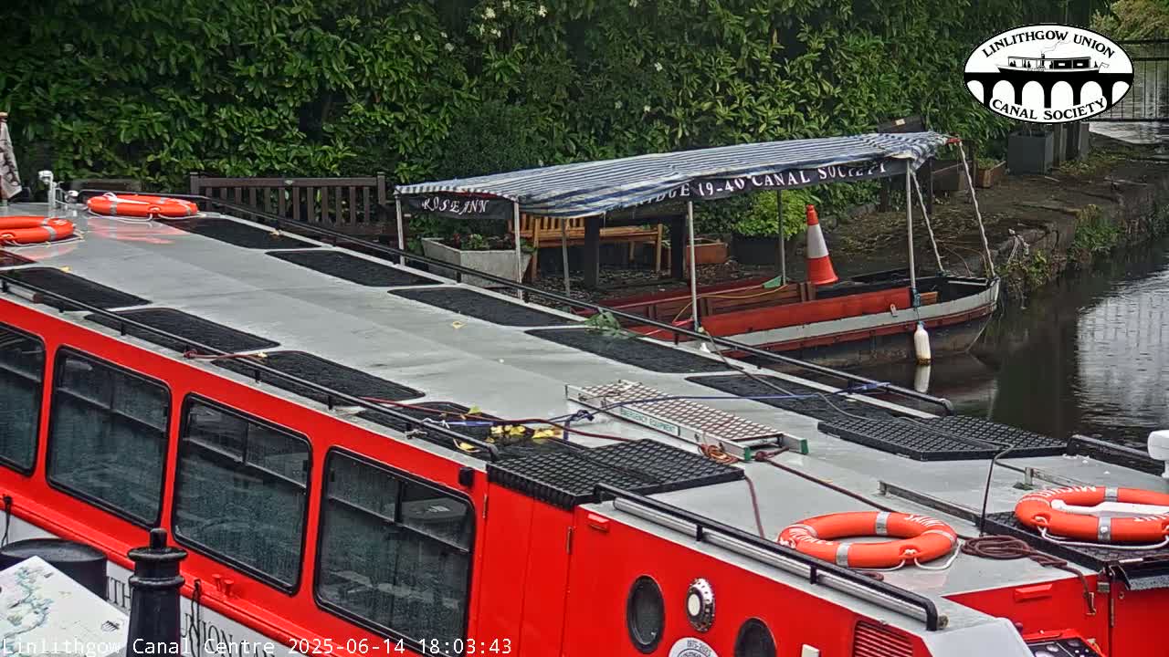 A red boat with life preservers on its roof is docked next to a smaller, covered boat on a calm canal under an overcast sky.