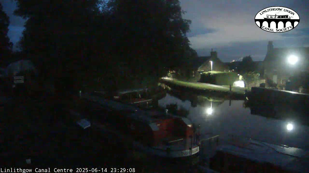 A red boat with life preservers on its roof is docked next to a smaller, covered boat on a calm canal under an overcast sky.