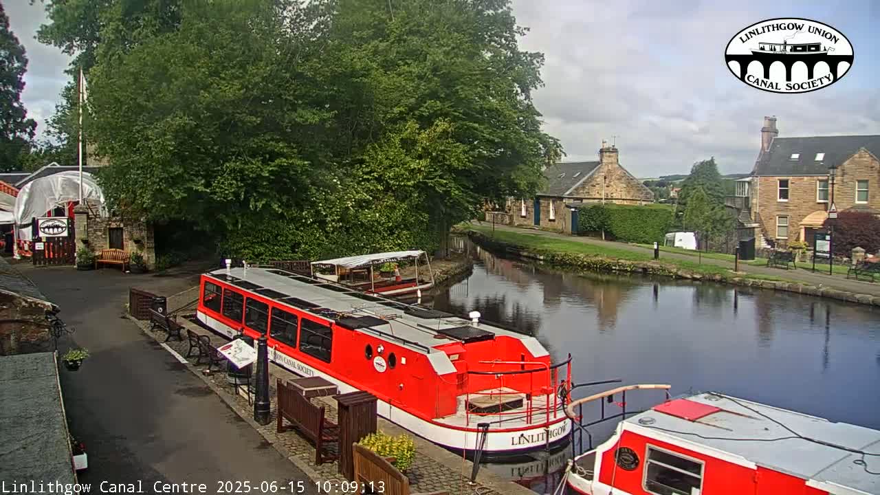 Two red canal boats are docked near a stone building beside a calm canal under a partly cloudy sky.