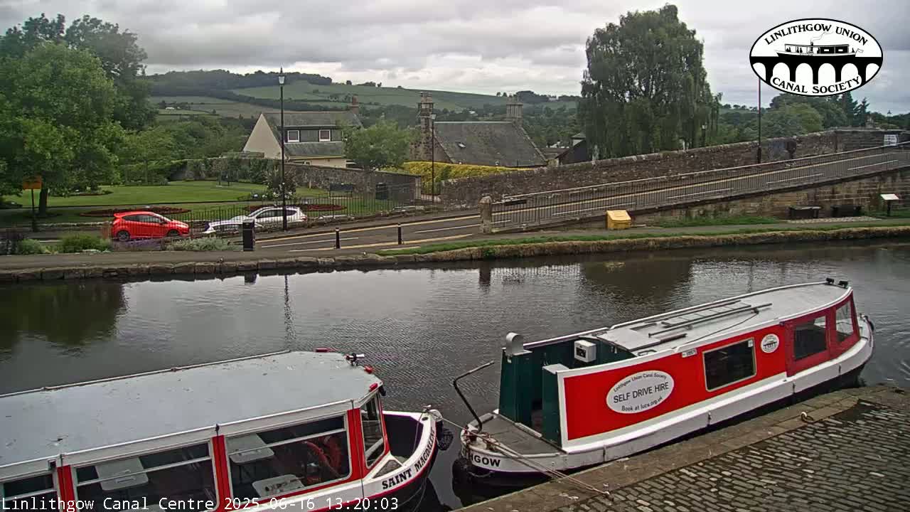 Two narrowboats are moored on a canal, next to a roadway and a stone bridge, under an overcast sky.