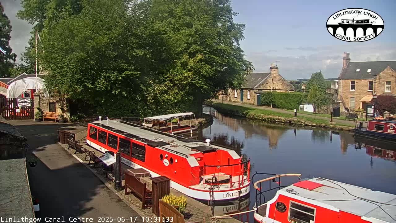 A sunny day shows two red canal boats moored near a stone building and lush green trees along a calm canal.