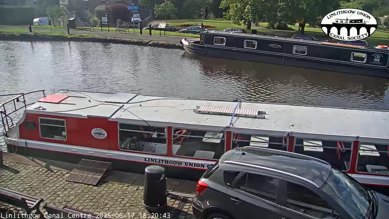 A red and white canal boat is docked next to a dark-colored SUV, with another black canal boat moored further down the waterway on a sunny day.