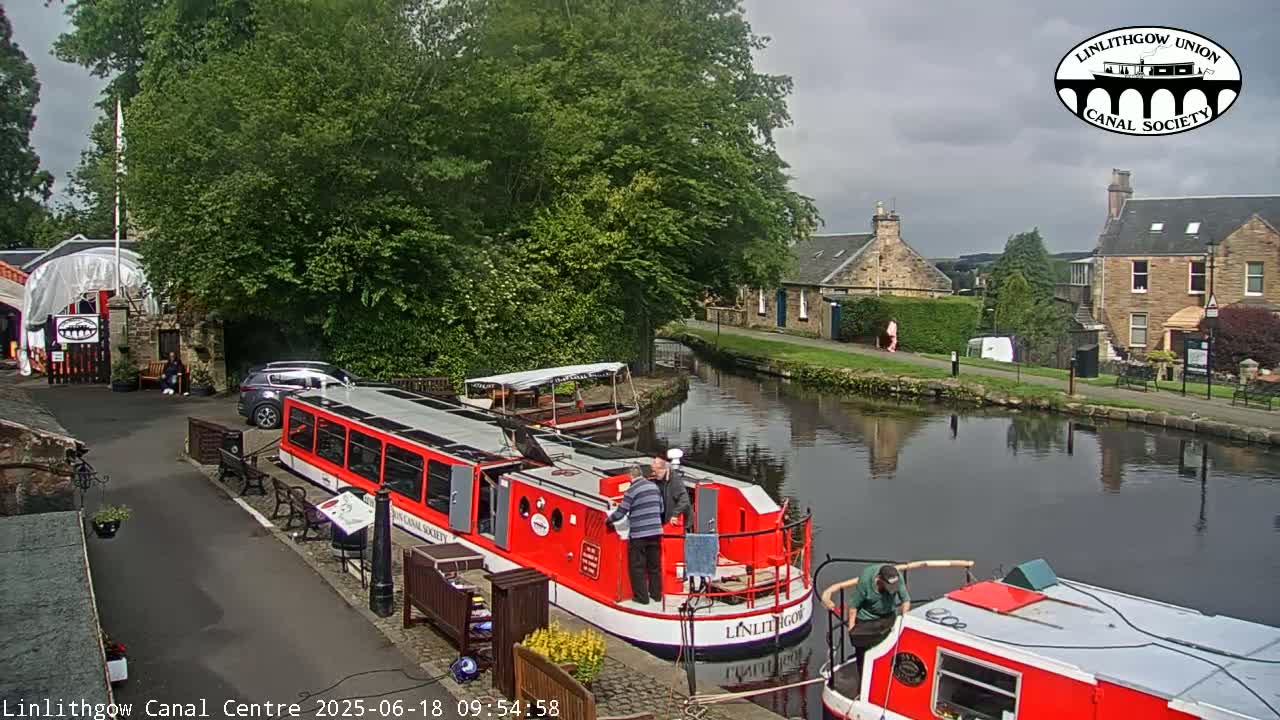 Two red canal boats are docked near a stone building beside a calm canal under a partly cloudy sky.