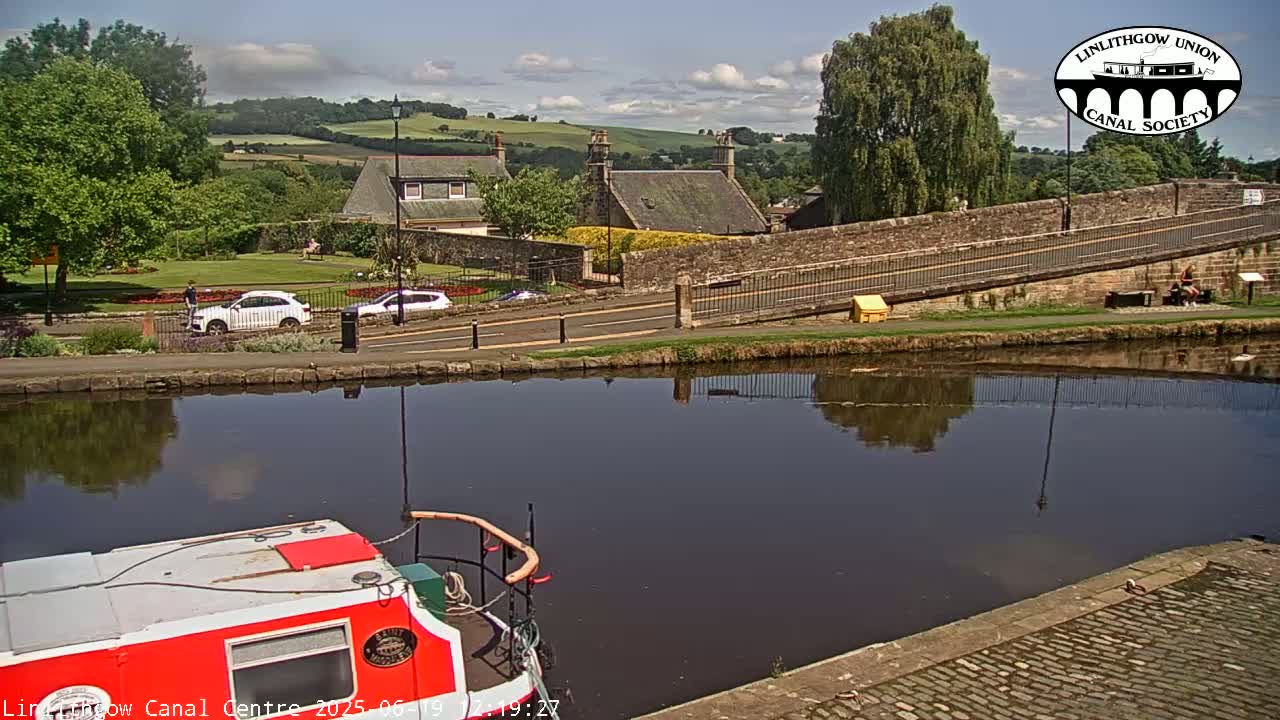 A red and white canal boat is moored beside a calm canal reflecting a stone bridge and buildings on a sunny day.
