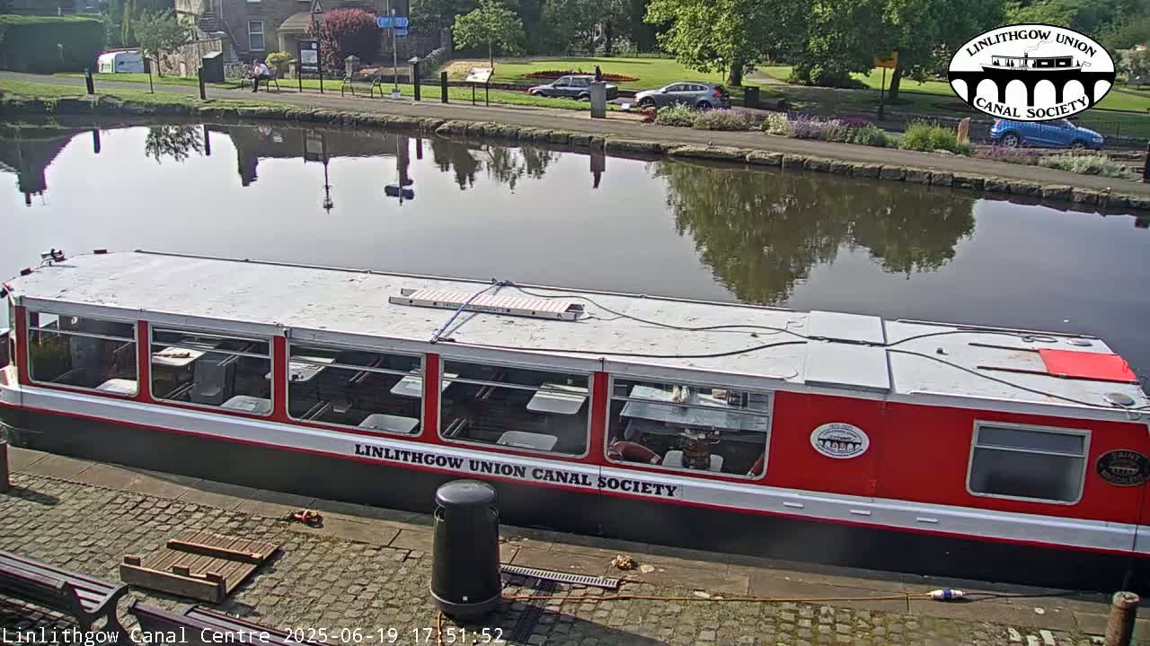 A red and white canal boat is docked at a canal, with calm water reflecting the surrounding trees and buildings on a sunny day.