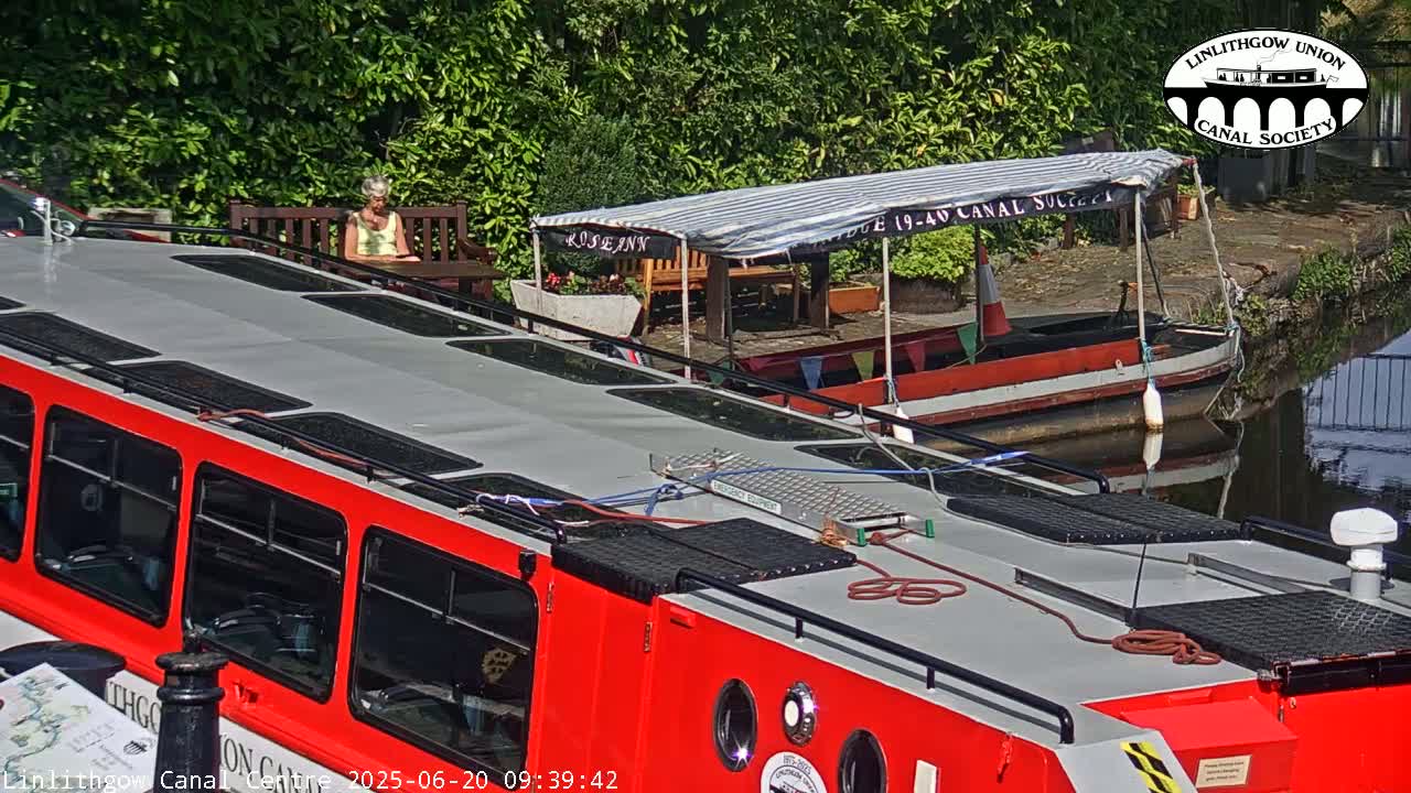 A red canal boat is moored beside a smaller, older boat, with a person sitting at a table under a striped awning on a sunny day.