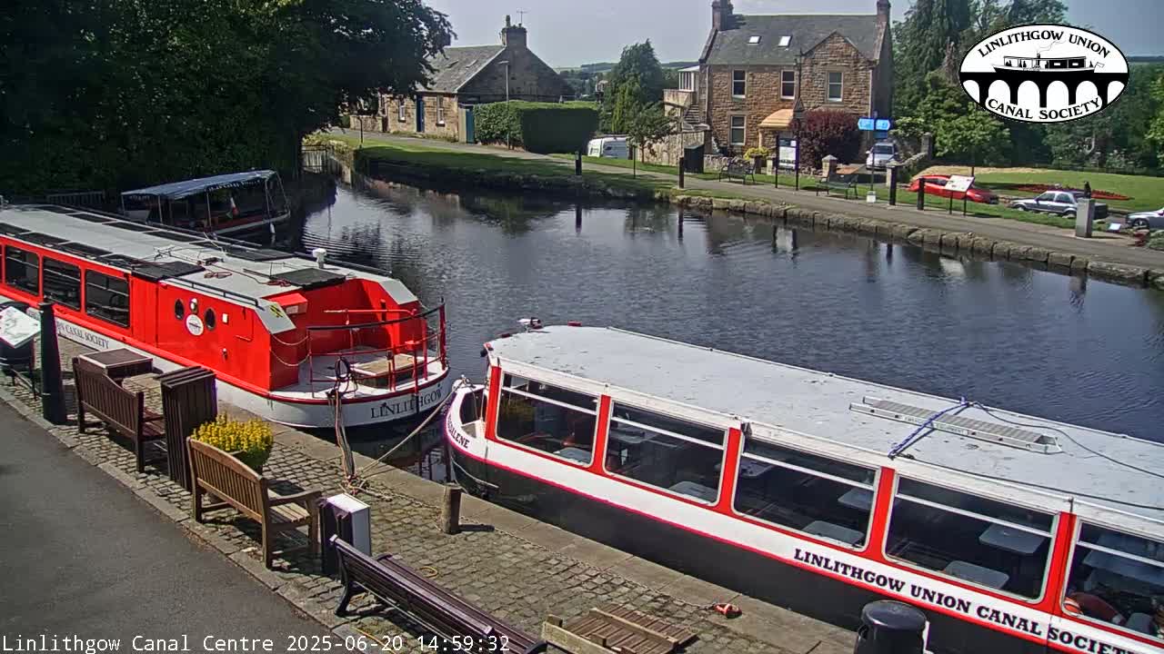 Two red and white canal boats are moored along a canal bank on a sunny day, with stone buildings and greenery visible in the background.