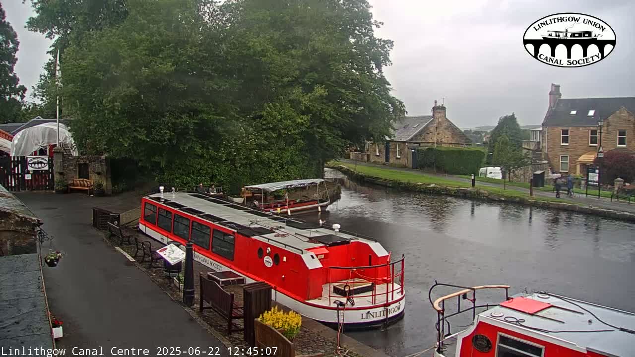A red canal boat is docked near a canal-side path, with another smaller boat moored further down the waterway under an overcast sky.