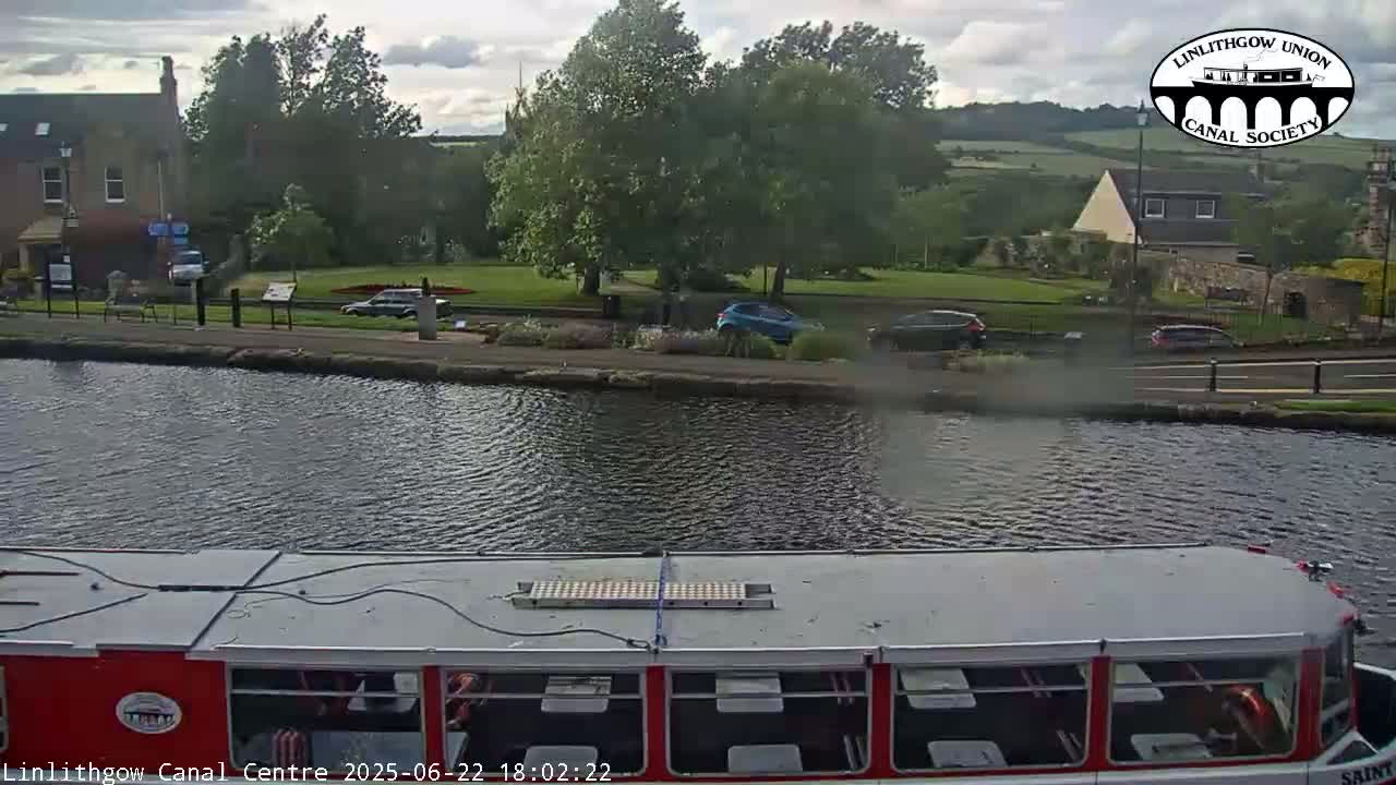 A red and white canal boat sits on the water, overlooking a canal-side path with cars parked along it and buildings and trees in the background under a partly cloudy sky.