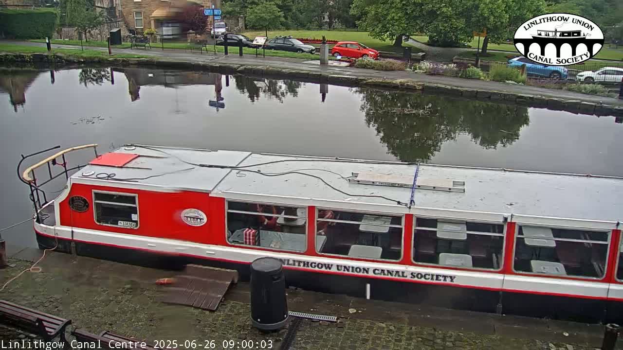 A red and white canal boat is docked on a calm, overcast day next to a paved walkway and several parked cars.