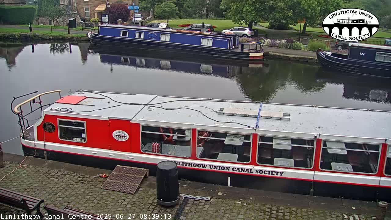 A red and white canal boat is docked next to a blue canal boat on a calm canal, with green trees and buildings in the background.