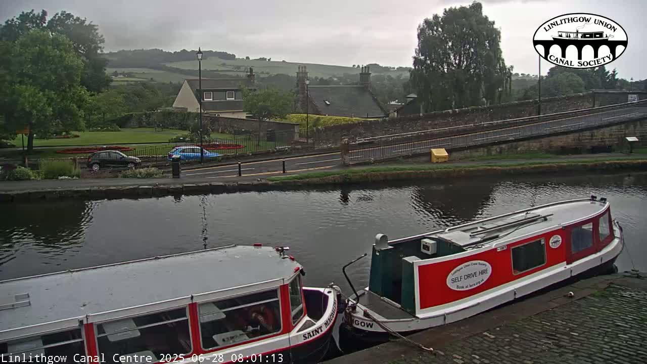 Two narrowboats are moored on a canal under an overcast sky, with a road and buildings visible in the background.