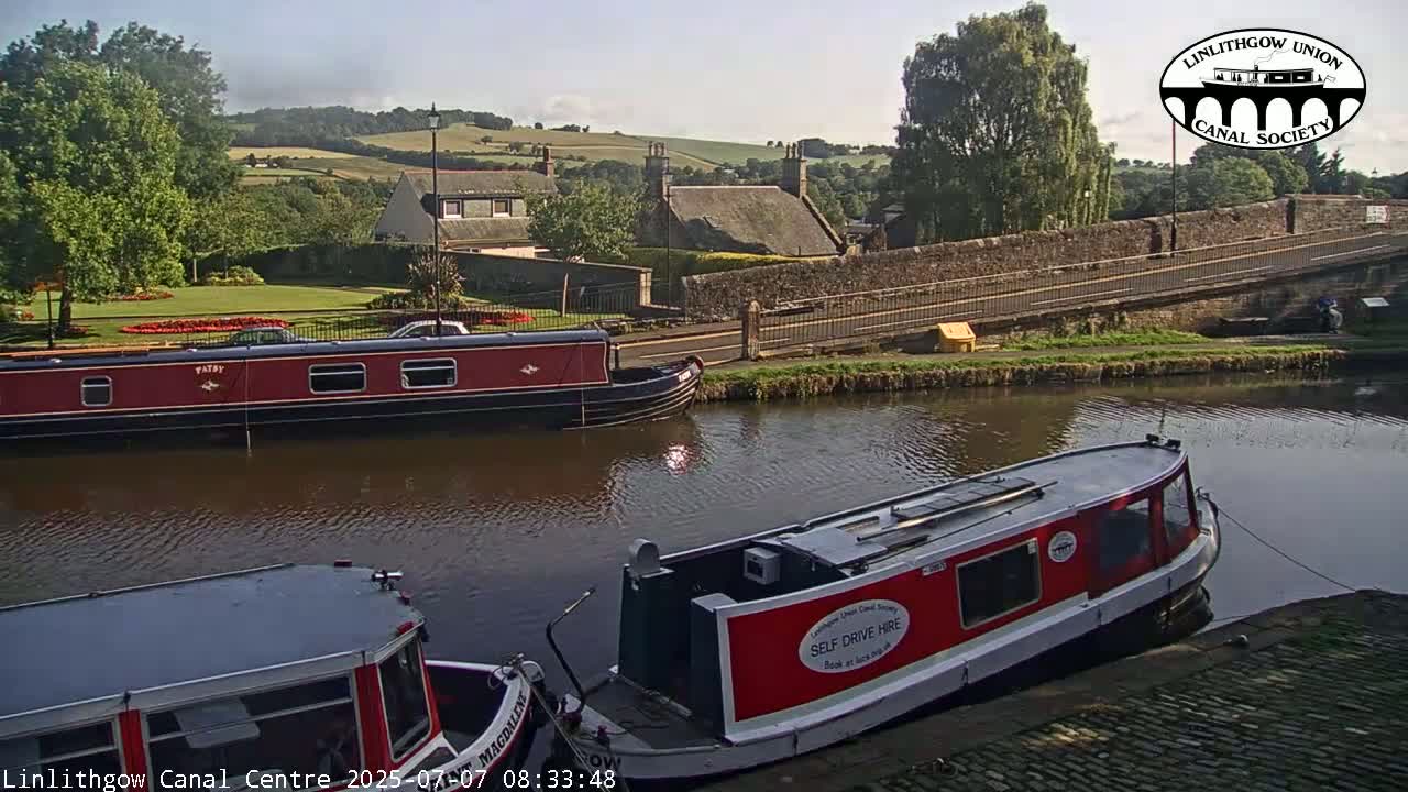 Two narrowboats are moored on a calm canal, with a stone bridge and distant houses visible under a sunny sky.