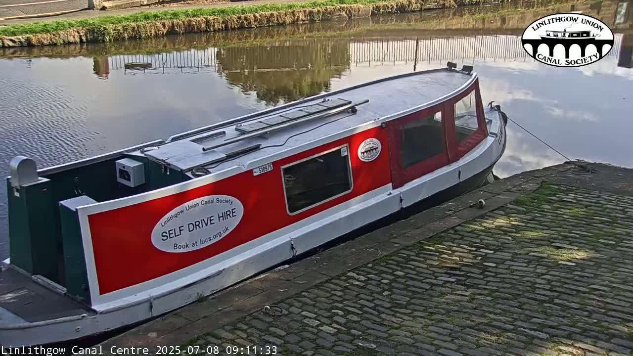 A red and white narrowboat is moored alongside a cobblestone quay beside a calm canal.
