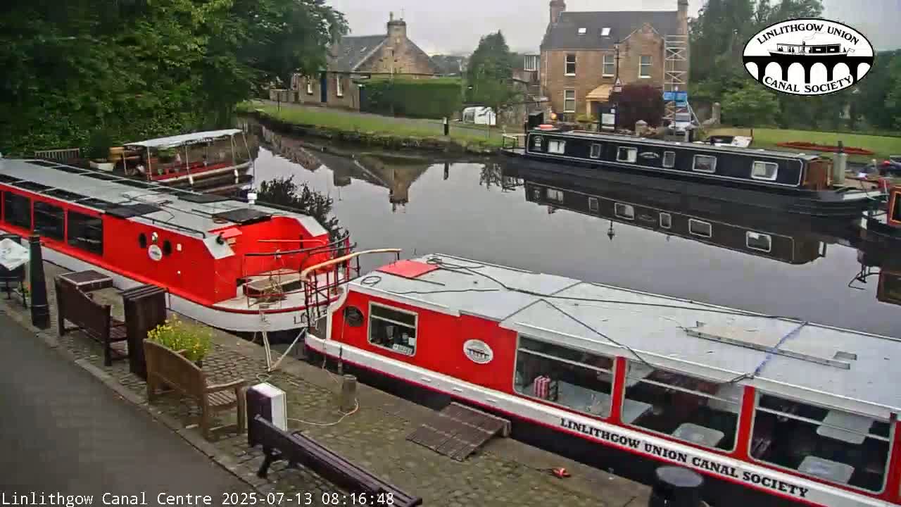 Two red canal boats and a black canal boat are moored on a calm canal, with buildings and trees visible in the background on an overcast day.