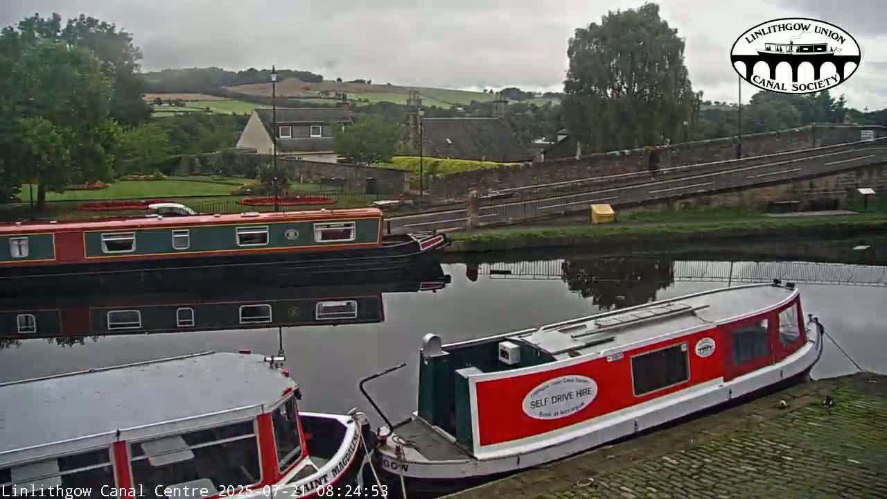 Two narrowboats are moored on a canal under an overcast sky, with a stone bridge and buildings visible in the background.