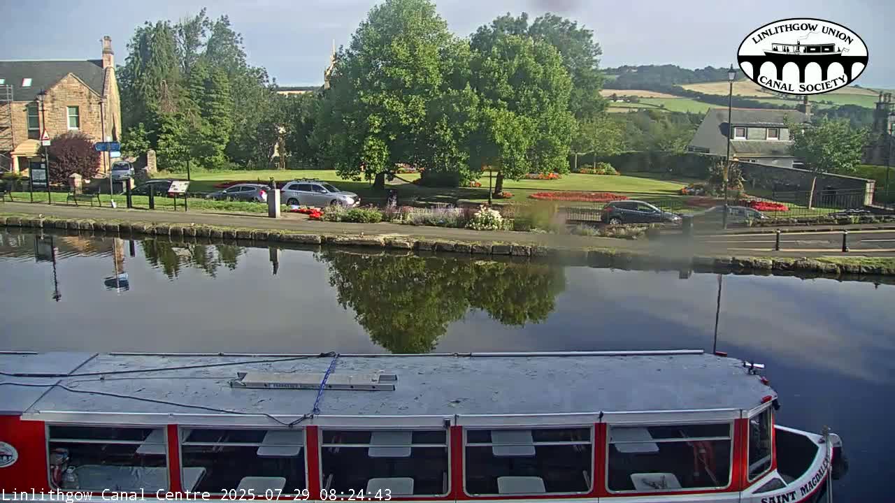 A red and gray canal boat is docked on a calm canal, reflecting buildings and lush greenery under a clear sky.