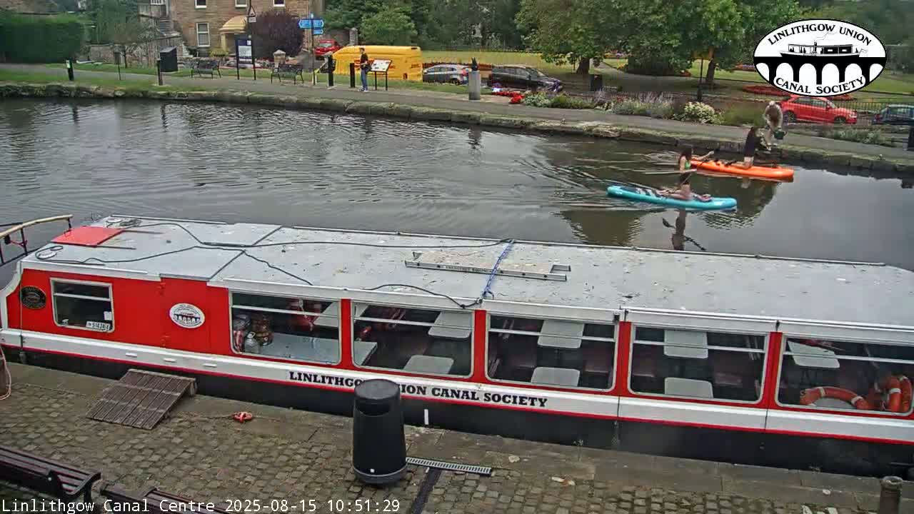 A red and gray canal boat is docked on a calm canal, reflecting buildings and lush greenery under a clear sky.