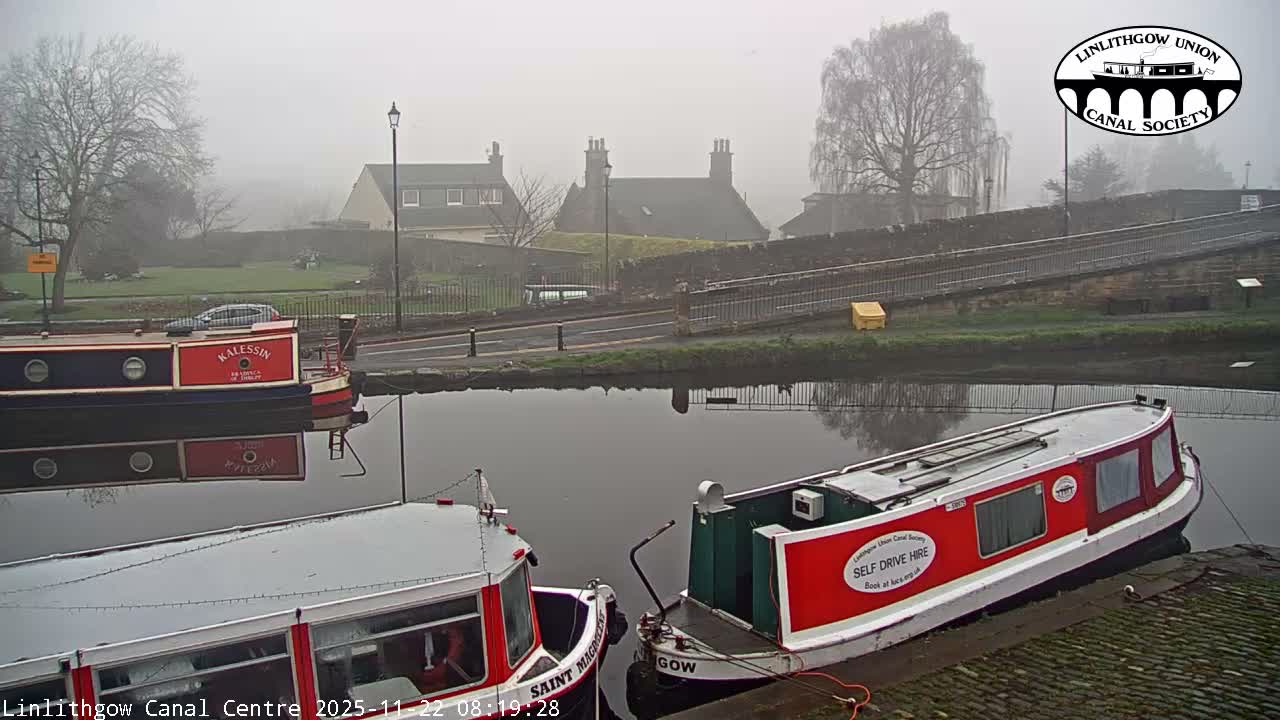 Three colorful canal boats are moored on a calm canal, with a road, houses, and bare trees shrouded in a thick fog in the background.