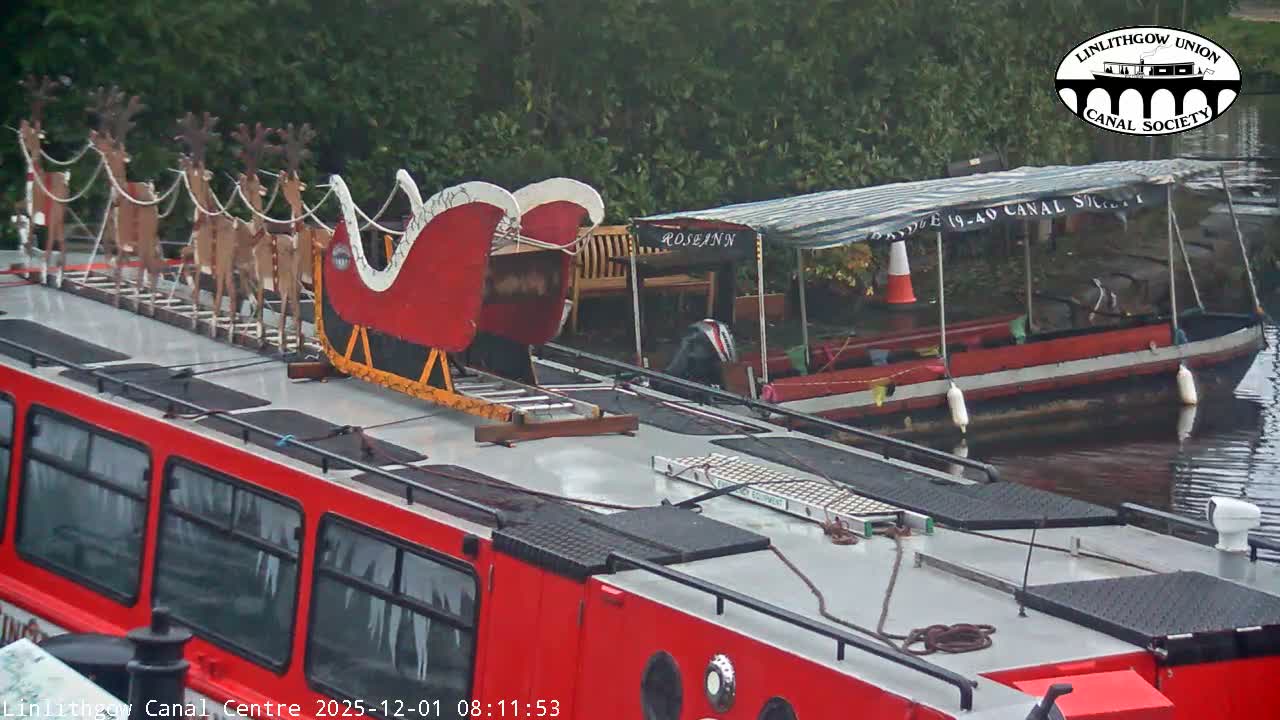 On an overcast day, a red canal boat adorned with a festive Santa's sleigh and reindeer figures is docked next to a smaller boat with a striped canopy, against a backdrop of green foliage.