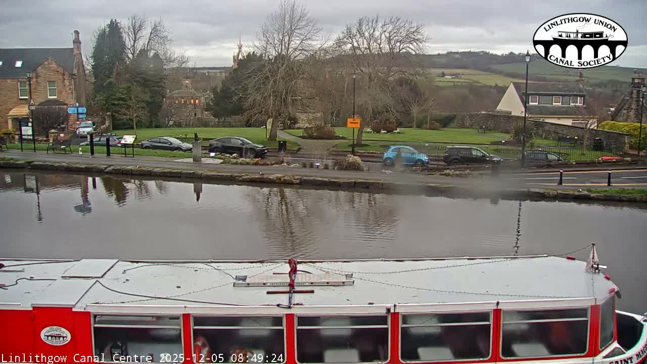 A canal boat fills the bottom of the frame, overlooking a tranquil canal with reflections, a path lined with parked cars and bare trees, and distant buildings and rolling hills under an overcast sky.