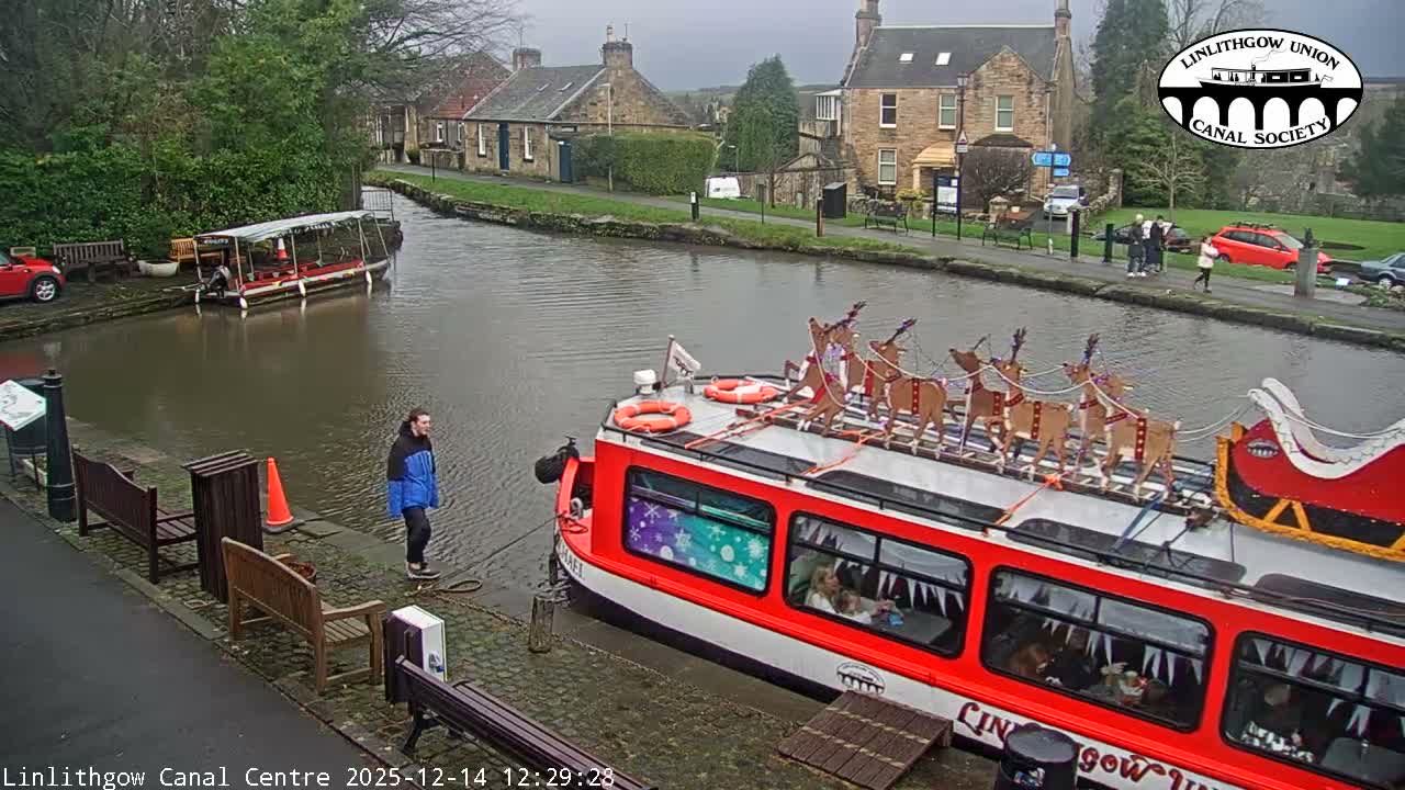 A canal boat fills the bottom of the frame, overlooking a tranquil canal with reflections, a path lined with parked cars and bare trees, and distant buildings and rolling hills under an overcast sky.