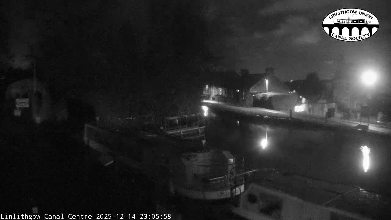 A canal boat fills the bottom of the frame, overlooking a tranquil canal with reflections, a path lined with parked cars and bare trees, and distant buildings and rolling hills under an overcast sky.