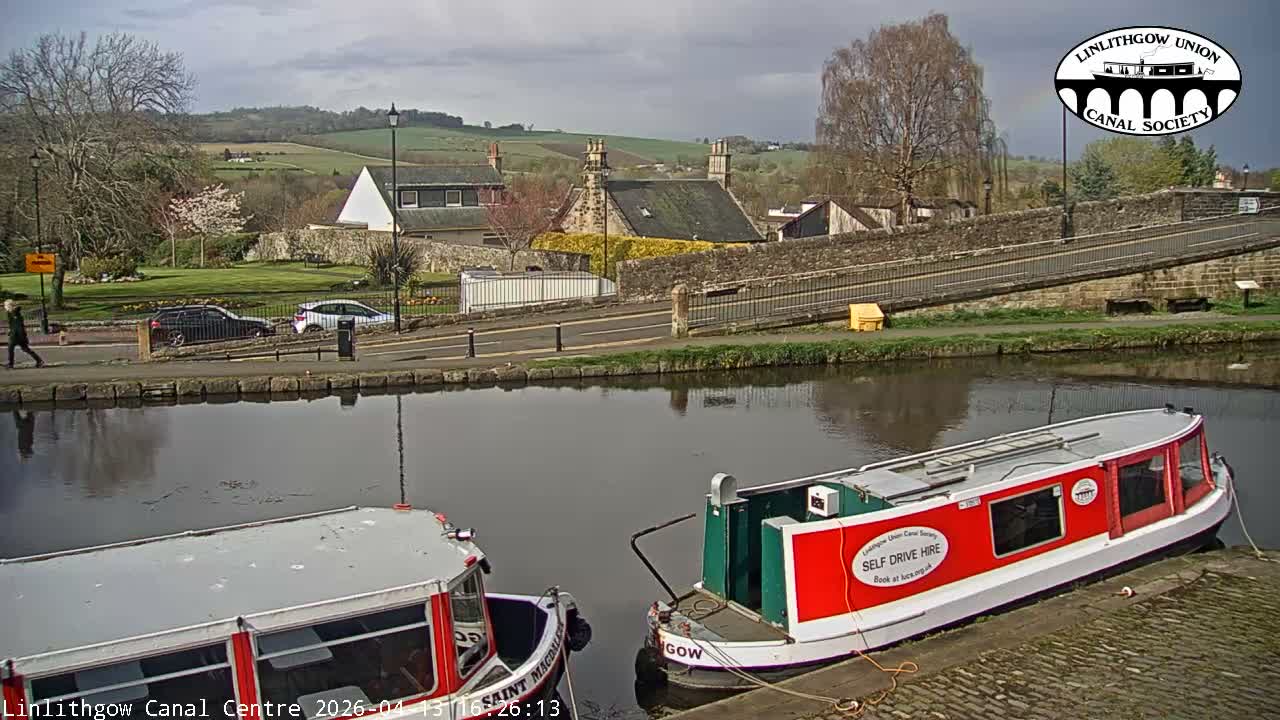 A canal boat fills the bottom of the frame, overlooking a tranquil canal with reflections, a path lined with parked cars and bare trees, and distant buildings and rolling hills under an overcast sky.