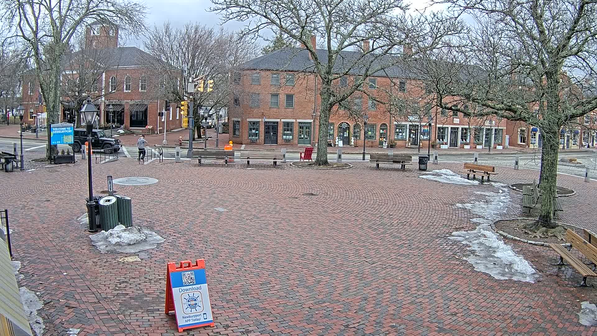 A brick plaza with benches and trees, partially covered in melting snow, sits in front of brick buildings on a cloudy day.