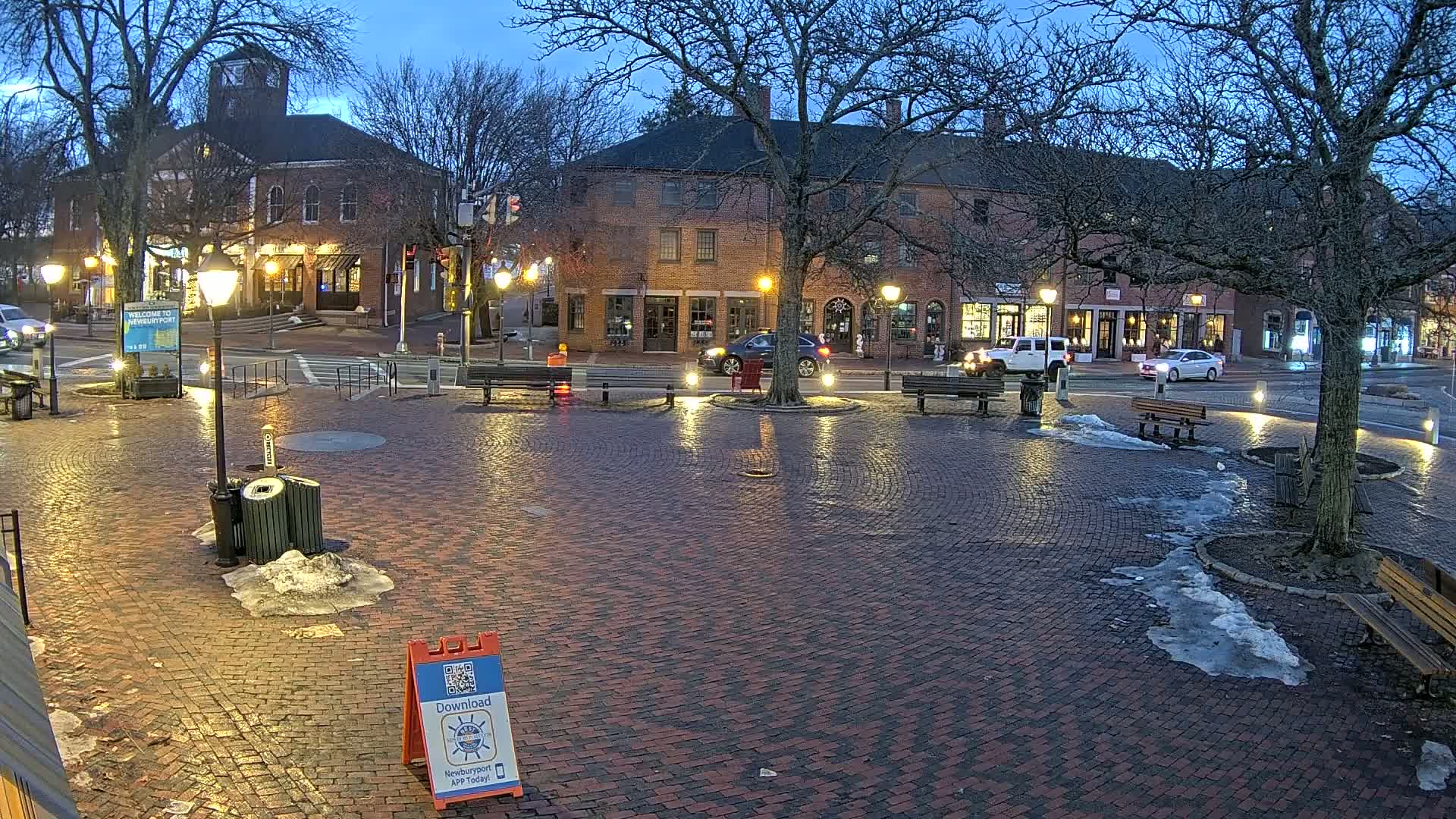A brick town square at twilight, partially covered in melting snow, is illuminated by streetlights, with several benches and cars parked nearby.