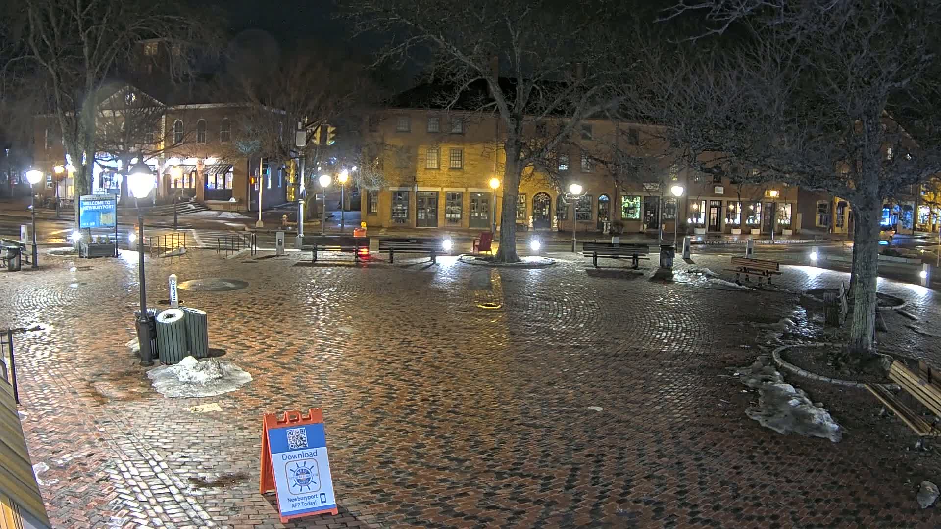 A brick plaza at night, under partly cloudy skies, is lit by streetlamps and shows patches of snow and ice on the ground around trees and benches.